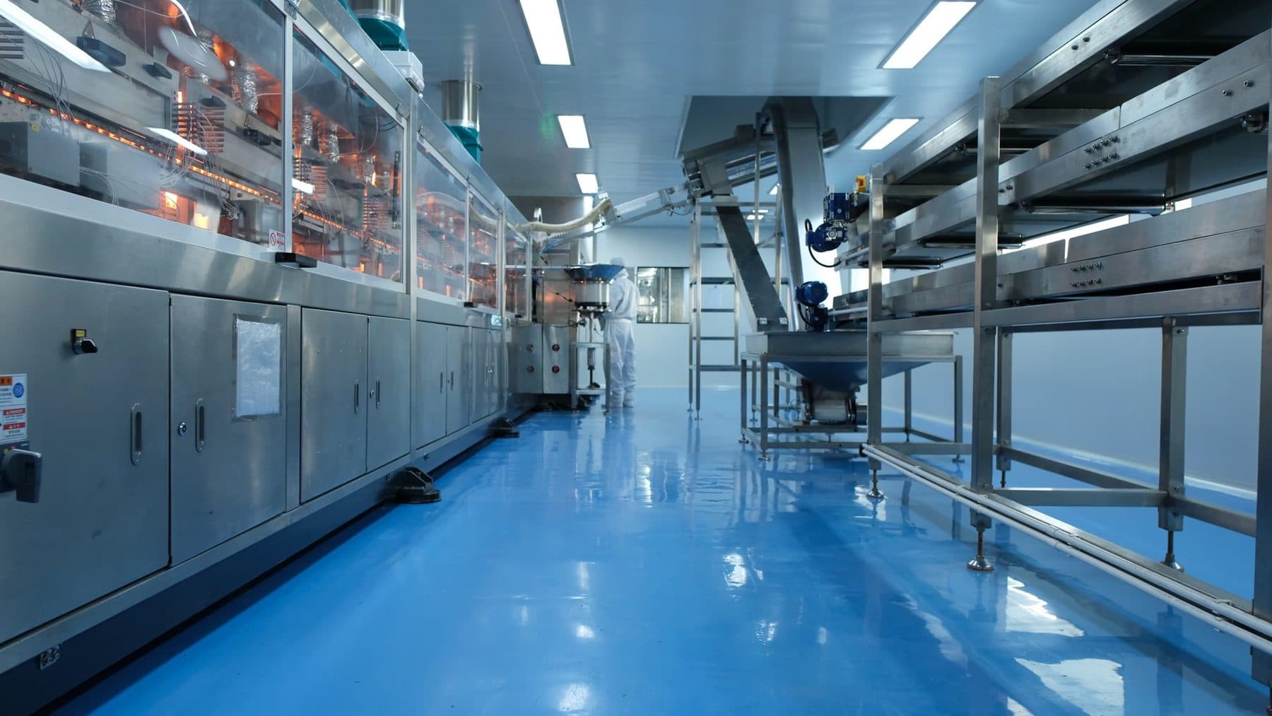 Inside the production hall at Satoria Pharma's Pasuruan plant: stainless-steel processing machinery and conveyors in a cleanroom, with an operator in full protective garments